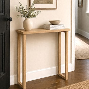 May include: A light wood console table with a rectangular top and four straight legs. On the table sits a beige ceramic vase containing green leafy branches, a small tan bowl, and two stacked white books titled Live Beautifully and This Is Home.