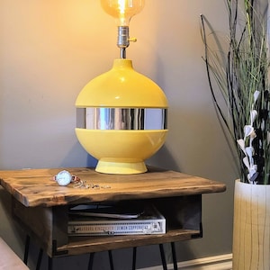 May include: A yellow mid-century modern lamp with a silver band sits on a wooden nightstand with a book and a watch. The nightstand has black hairpin legs. A vase with dried flowers is in the background.
