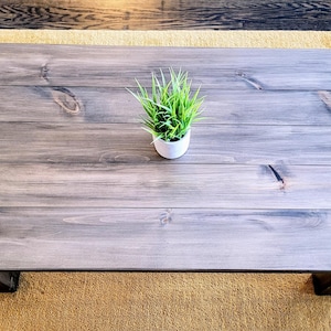 Peut inclure: Une table basse en bois gris avec une teinte gris clair. La table a un design simple avec un plateau rectangulaire et quatre pieds. Une petite plante en pot est placée au centre de la table.