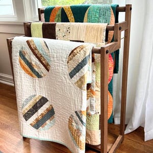 May include: A wooden quilt rack holding three folded fabric quilts. The front quilt is cream-coloured with large circular patterns featuring brown, blue, and tan stripes. The middle quilt has brown and floral panels, and the back quilt is teal.