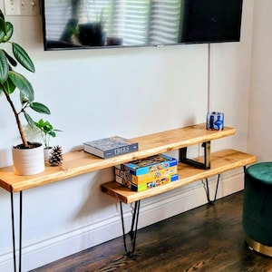 May include: A two-tiered, natural wood TV stand with black hairpin legs. The top shelf holds a book and a small pinecone, while the lower shelf displays a colorful box. A potted plant and a green ottoman are also visible.