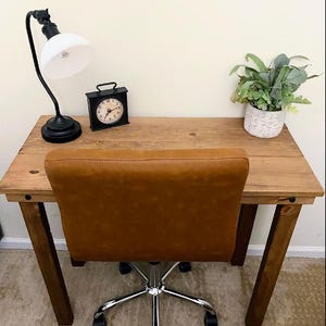 May include: A home office setup with a wooden desk, a brown leather office chair, a black desk lamp, a small black clock, and a potted plant. The desk has a natural wood finish, and the chair has a chrome base.