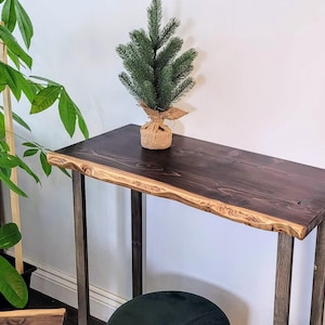 Peut inclure: Une console en bois de bord brut de couleur brun foncé avec un cadre en métal. La table a un grain de bois naturel et une finition rustique. Un petit arbre de Noël en pot est posé sur la table.