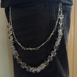 May include: A silver-toned, double-strand chain with clear, faceted beads and wire detailing. The chain is attached to a black garment, likely jeans. The design incorporates a barbed wire element, adding a unique, edgy aesthetic.