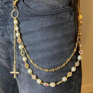 May include: Gold-tone chain with a lobster clasp, attached to a pair of black jeans. The chain features faux pearls, colorful gemstones, and a cross pendant. A second chain with star and bead accents hangs from the jeans.