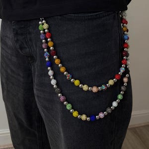 May include: A multi-colored beaded chain with silver-tone hardware hangs from a person's pocket. The chain features a variety of colorful beads, including red, yellow, blue, green, and purple. The person is wearing black jeans and a black shirt.
