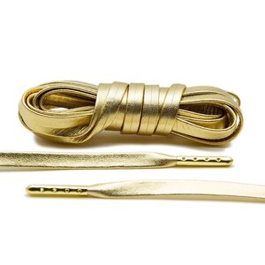 May include: Gold leather shoelaces with a metallic finish. The laces are tied in a knot and laid out flat on a white background.