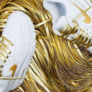 May include: White leather sneakers with gold laces and a gold Nike swoosh logo. The sneakers are surrounded by gold ribbon.