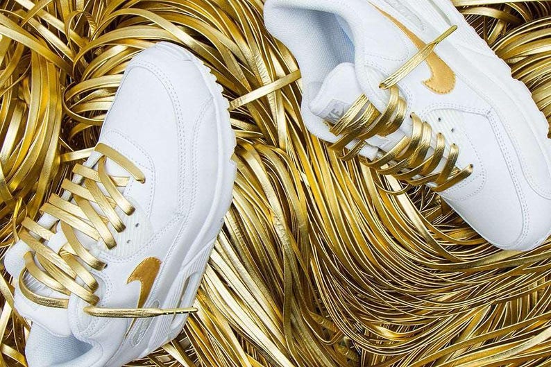 May include: White leather sneakers with gold laces and a gold Nike swoosh logo. The sneakers are surrounded by gold ribbon.
