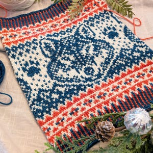 Könnte beinhalten: Ein unfertiges Strickprojekt mit einem komplexen Muster in Marineblau, Weiß und Orange. Das Design zeigt geometrische und stilisierte Blumenmotive. Ein Knäuel weiße Wolle und Grünzeug befinden sich im Hintergrund.
