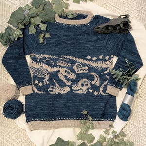 以下が含まれることがあります： 胸にベージュの恐竜の骨格模様が入った青いニットセーター。セーターはベージュの襟、袖口、裾が付いています。青い糸の束とベージュの糸玉がセーターの隣にあります。