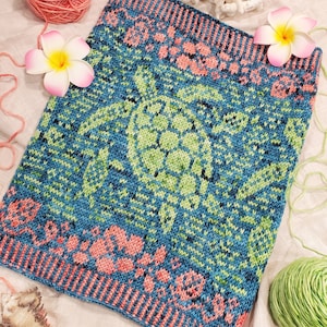 Puede incluir: Una bufanda de punto azul y verde con un diseño de tortuga marina y detalles florales rosas. La bufanda está hecha con un hilo texturizado y tiene un borde con flecos.