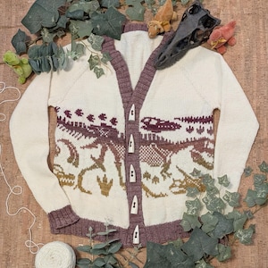 May include: Cream-coloured knitted cardigan featuring a dinosaur design in shades of brown and gold. The cardigan has a V-neck, long sleeves, and a button-up front. The trim is a deep burgundy colour.