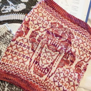 May include: A knitted cowl in shades of red, pink, and cream, featuring a geometric pattern with tree-like motifs. The cowl is displayed on top of an open book, showcasing the intricate design and craftsmanship of the handmade item.
