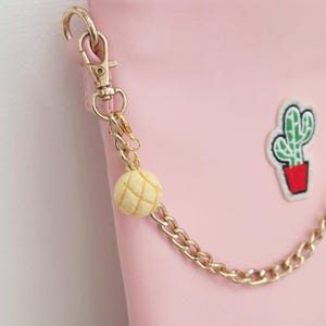 May include: A light pink pouch featuring a gold-toned metal chain strap attached with a lobster clasp. The chain is adorned with a small, round, textured beige charm resembling a bun and a small embroidered patch of a green cactus in a red pot.