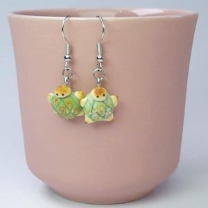 Puede incluir: Un par de pendientes de plata con colgantes de tortugas verdes y marrones. Las tortugas tienen un caparazón texturizado y una pequeña cara.