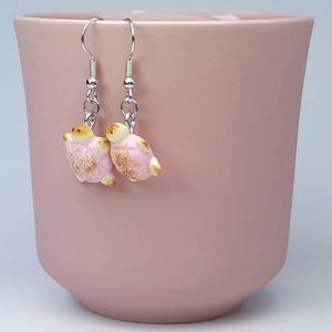 Op de afbeelding: Een paar roze en gouden schildpad oorbellen met zilveren haakjes. De oorbellen hangen aan een roze kopje.