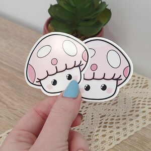 Op de afbeelding: Twee gestanste stickers met een cartoon paddenstoelkarakter met een lichtroze hoed, witte stippen en een lachend gezicht met roze wangen. De stickers hebben een glanzende afwerking en een witte rand voor een strak grafisch ontwerp.