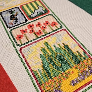 May include: A cross-stitch design featuring scenes from The Wizard of Oz. The design includes a hot air balloon, the yellow brick road, the Wicked Witch, poppies, and the Emerald City, all framed in green.