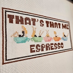 May include: Cross-stitch embroidery with brown thread on white fabric. The embroidery says "That's That Me Espresso" with a row of five colorful cups below the text.