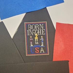 May include: A cross-stitched patch with the text "BORN IN THE USA" and three figures on a podium. The patch is framed in red and blue, set against a black fabric, with blue, white, and red felt pieces in the background.