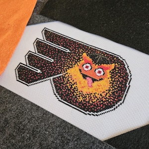 May include: Cross-stitch design featuring a cartoonish orange and yellow creature with a black hat and a pink tongue, set against a black and red speckled background. The design is on a white fabric with a black outline.