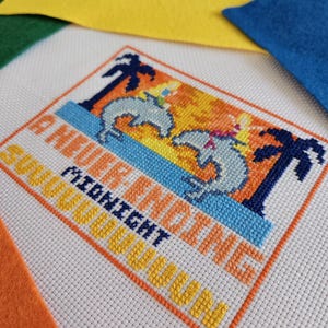 Puede incluir: Obra de arte de punto de cruz que representa una escena tropical con delfines, palmeras y el texto "A Never Ending Midnight Moon". El diseño utiliza hilo azul, naranja, amarillo y blanco sobre lienzo blanco. Cuadrados de fieltro en amarillo, azul y naranja en el fondo.