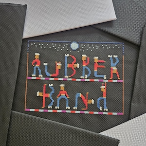 以下が含まれることがあります： 黒い布に施されたクロスステッチ刺繍で、RUBBER BANDという文字が描かれています。各文字は赤と青の服を着たデフォルメされた人物で構成されています。デザインには小さな月と星が含まれ、下部にはチェック柄の縁取りが施されています。