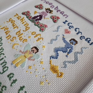 May include: Cross-stitch artwork in a white frame. The design features whimsical figures, flowers, and text that reads "moonbeam ice cream" and "taking your blue jeans off". The color palette includes blue, yellow, green, and pink.