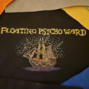 May include: A black canvas with gold embroidery depicting a ship sailing under a starry sky. The text "Floating Psycho Ward" is embroidered above the ship.