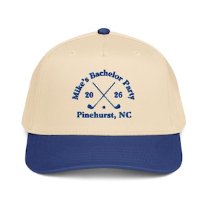 May include: A baseball cap with a cream-colored crown and a blue brim. The cap features the text "Mike's Bachelor Party" with crossed golf clubs and the year "20 26" in blue, along with "Pinehurst, NC".