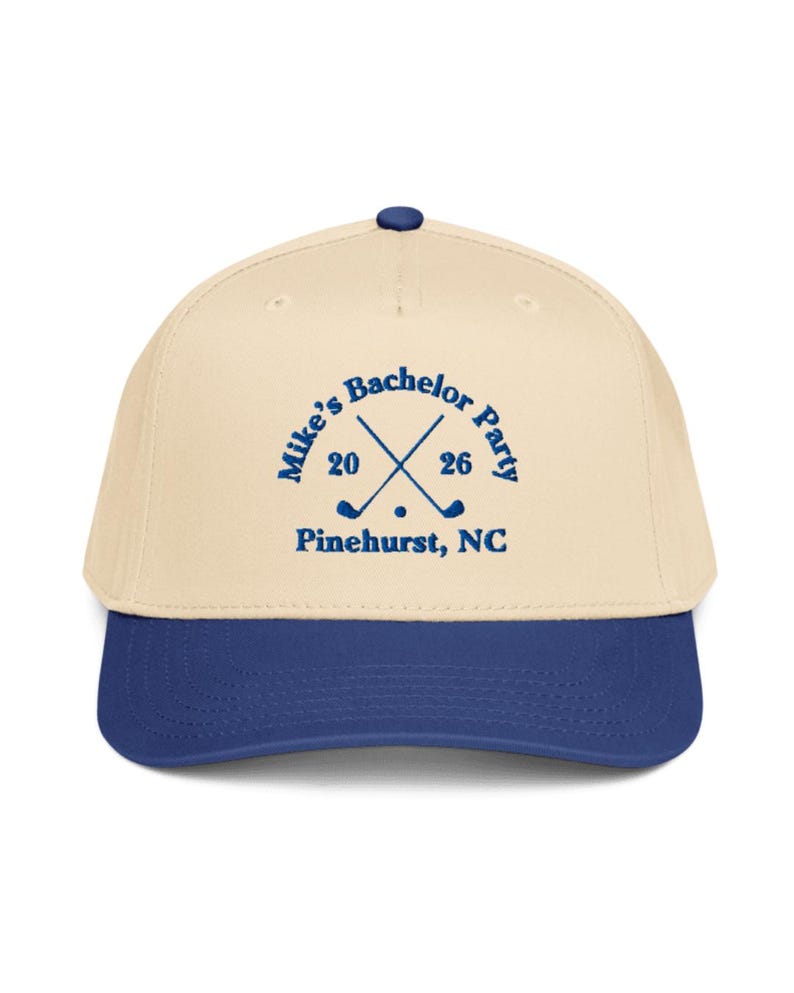 May include: A baseball cap with a cream-colored crown and a blue brim. The cap features the text "Mike's Bachelor Party" with crossed golf clubs and the year "20 26" in blue, along with "Pinehurst, NC".