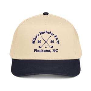 May include: A beige and navy blue baseball cap with the text "Mike's Bachelor Party 2026" and crossed golf clubs. The text "Pinehurst, NC" is printed below the crossed golf clubs. The cap has a curved brim and a button on top.