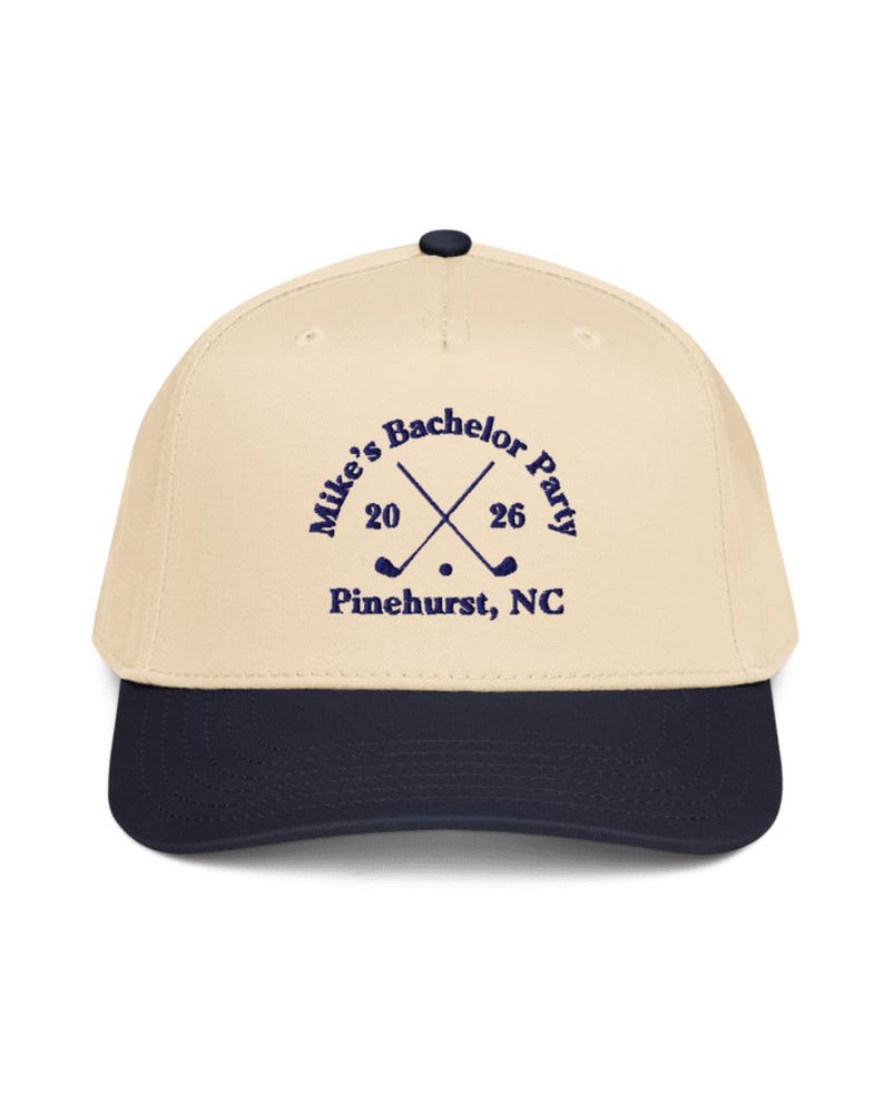 May include: A beige and navy blue baseball cap with the text "Mike's Bachelor Party 2026" and crossed golf clubs. The text "Pinehurst, NC" is printed below the crossed golf clubs. The cap has a curved brim and a button on top.
