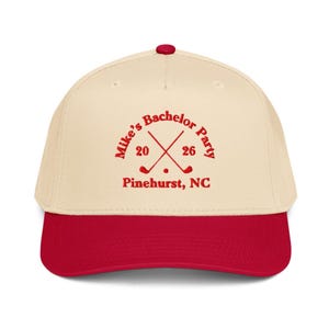 May include: A cream-colored baseball cap with a red brim and button. The cap features the text "Mike's Bachelor Party" in red, along with crossed golf clubs and the numbers "20" and "26". Below the text is "Pinehurst, NC".