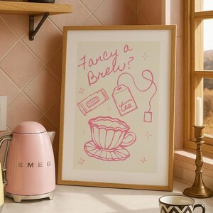以下が含まれることがあります： 「Fancy a Brew?」の文字と、ティーカップ、ティーバッグ、砂糖のイラストが描かれたピンク色のフレーム入りプリント。ピンク色のSMEGケトルと黄色のトースターがカウンターにあります。白黒模様のティーカップとソーサーも存在します。