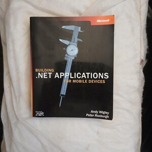 Peut inclure: Un livre intitulé Building .NET Applications for Mobile Devices par Andy Wigley et Peter Roxburgh. La couverture noire présente un graphique de pied à coulisse argenté, le logo Microsoft et le logo .NET avec du texte blanc et orange.