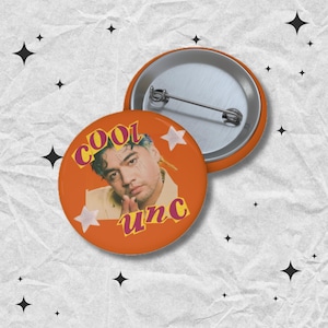 May include: Orange button with a portrait of a person with blue and yellow hair, the word "COOL" in pink, and the word "unc" in yellow. Two white star shapes are also on the button. A second button is visible.