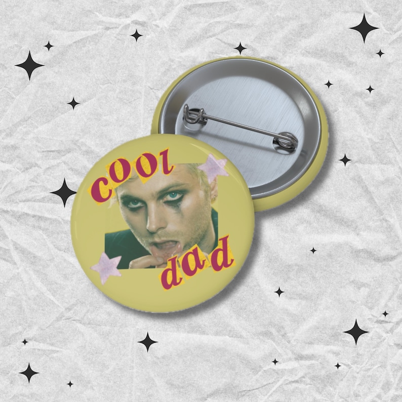 May include: A yellow button with the words "cool dad" in pink and a picture of a person with dark eye makeup. The button is round and has a pin on the back. Two pink star shapes are on the button.