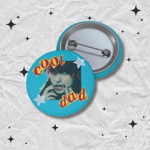 May include: Two round, teal-colored buttons. One button features an image of a person with red hair and the words "cool dad" in yellow and red text. The other button is silver and teal. White star shapes are also on the button.