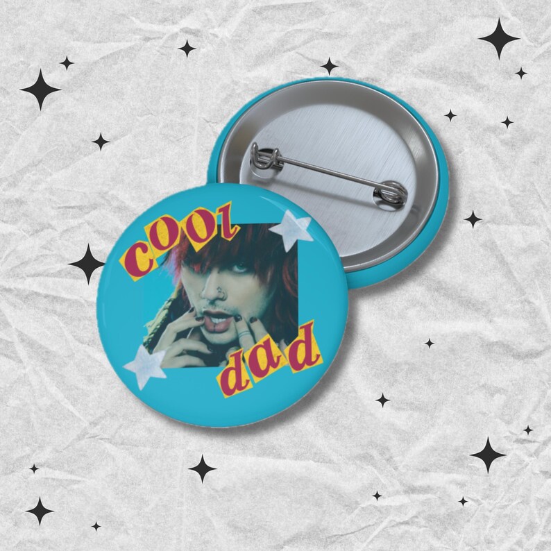 May include: Two round, teal-colored buttons. One button features an image of a person with red hair and the words "cool dad" in yellow and red text. The other button is silver and teal. White star shapes are also on the button.