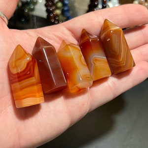 Puede incluir: Cinco puntas de cristal de cornalina, cada una con un patrón de bandas único de naranja, rojo y blanco. Las puntas están dispuestas en una fila, con la punta más grande a la derecha y la punta más pequeña a la izquierda.