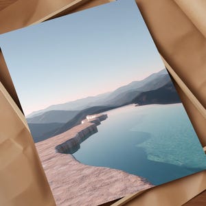 May include: A framed print featuring a serene landscape with a turquoise pool and layered mountains under a clear blue sky. The print is set in a light wooden frame, resting on brown paper.