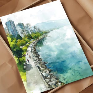 May include: Watercolour painting of a coastal city scene. The artwork depicts a waterfront promenade with cyclists, buildings, and a mountain range in the background. The painting is framed in a wooden box and set on a brown paper surface.