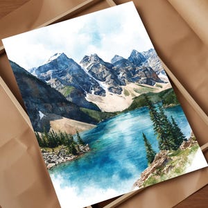 May include: Watercolor print of a mountain landscape with a turquoise lake and evergreen trees. The snow-capped mountains are in the background, and the artwork is framed in a wooden box, resting on brown paper.