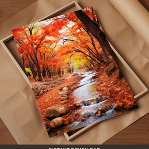 Peut inclure: Une impression encadrée représentant un paysage automnal. L'œuvre d'art montre un sentier bordant un ruisseau, entouré d'arbres aux feuilles rouge et orange flamboyantes. L'impression est dans un cadre en bois clair, posée sur une surface brune.