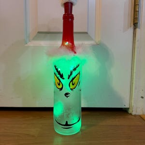 Puede incluir: Botella de vino decorativa con diseño de la cara del Grinch. La botella tiene ojos amarillos y cara verde, tapón rojo y ribete de piel blanca. La botella está iluminada con luces verdes. Decoración navideña.