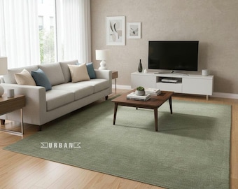 Puede incluir: Una alfombra rectangular verde salvia en un salón. La alfombra está colocada sobre un suelo de madera, con un sofá de color claro, una mesa de centro y un mueble de entretenimiento en el fondo. La palabra "URBAN" está impresa en la alfombra.