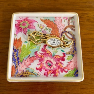 Peut inclure: Une montre-bracelet dorée avec un cadran blanc et des chiffres romains noirs repose sur un plateau carré à motifs floraux. Le plateau présente un fond blanc avec des motifs floraux roses, rouges, oranges et bleus, et une bordure dorée.