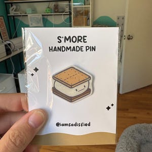 May include: A handmade S'more pin on a white card. The pin features a cartoon s'more with a graham cracker top and bottom, marshmallow filling, and a smiling face. The card reads "S'MORE HANDMADE PIN" and includes the Instagram handle @iamsatisfied.
